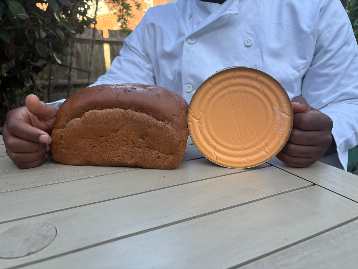 Person holding a loaf of spiced bun and a round tin of cheese  on a wooden surface outdoors.